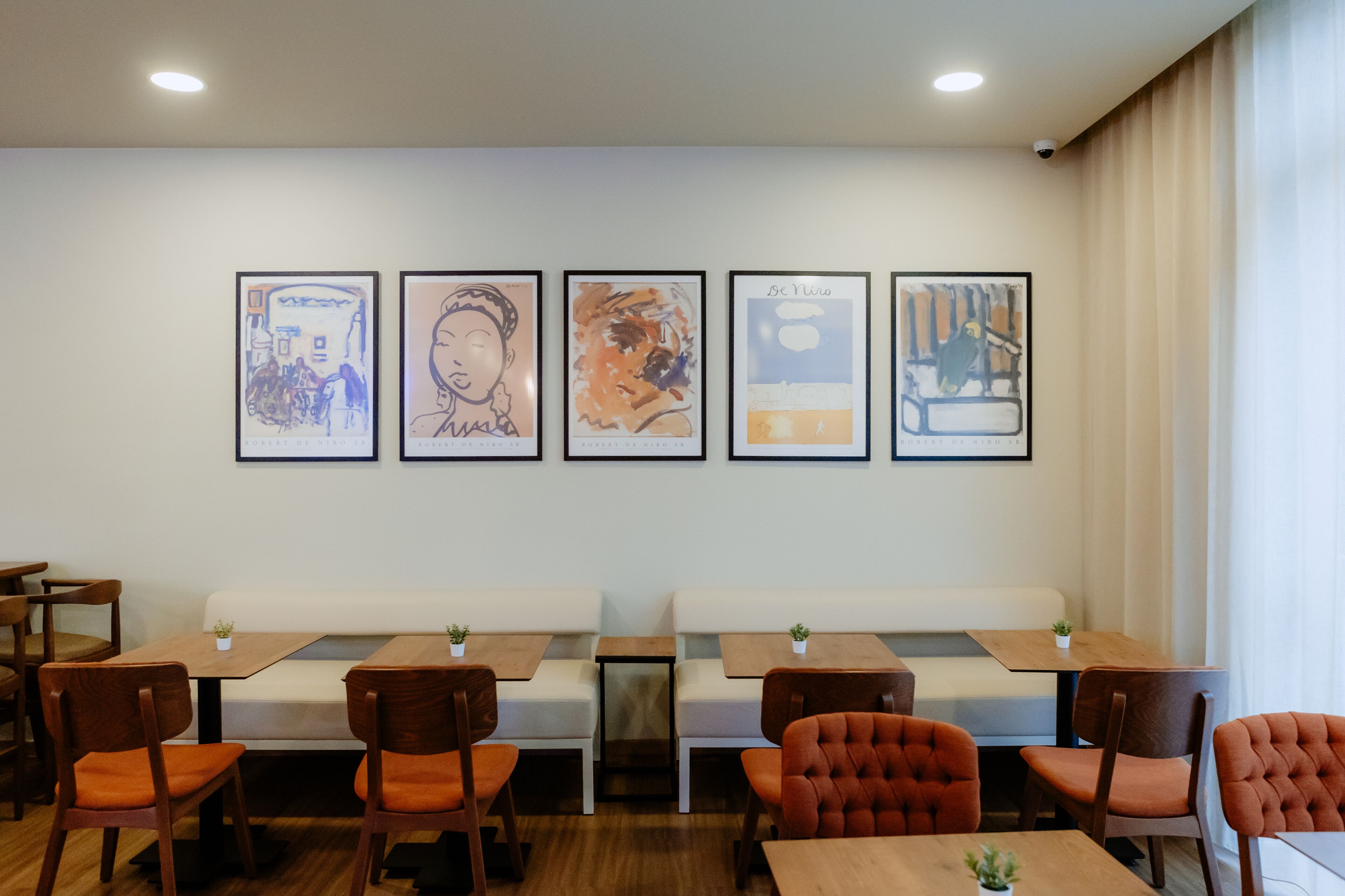 A restaurant interior with wooden tables and chairs, a white bench, and five framed artworks on the wall.