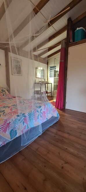 A bedroom with a bed covered by a mosquito net, wooden flooring, a mirror, and a wardrobe.