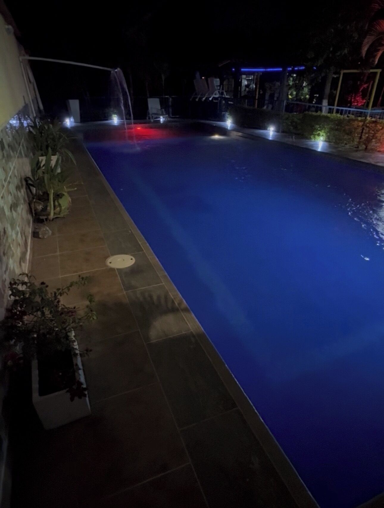 Una piscina nocturna con suelo de baldosas, algunas plantas en macetas y un edificio con entrada iluminada en azul.