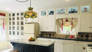 Una cocina con armarios blancos, una isla negra, una lámpara colgante con motivos florales y una ventana con cortinas decorativas.