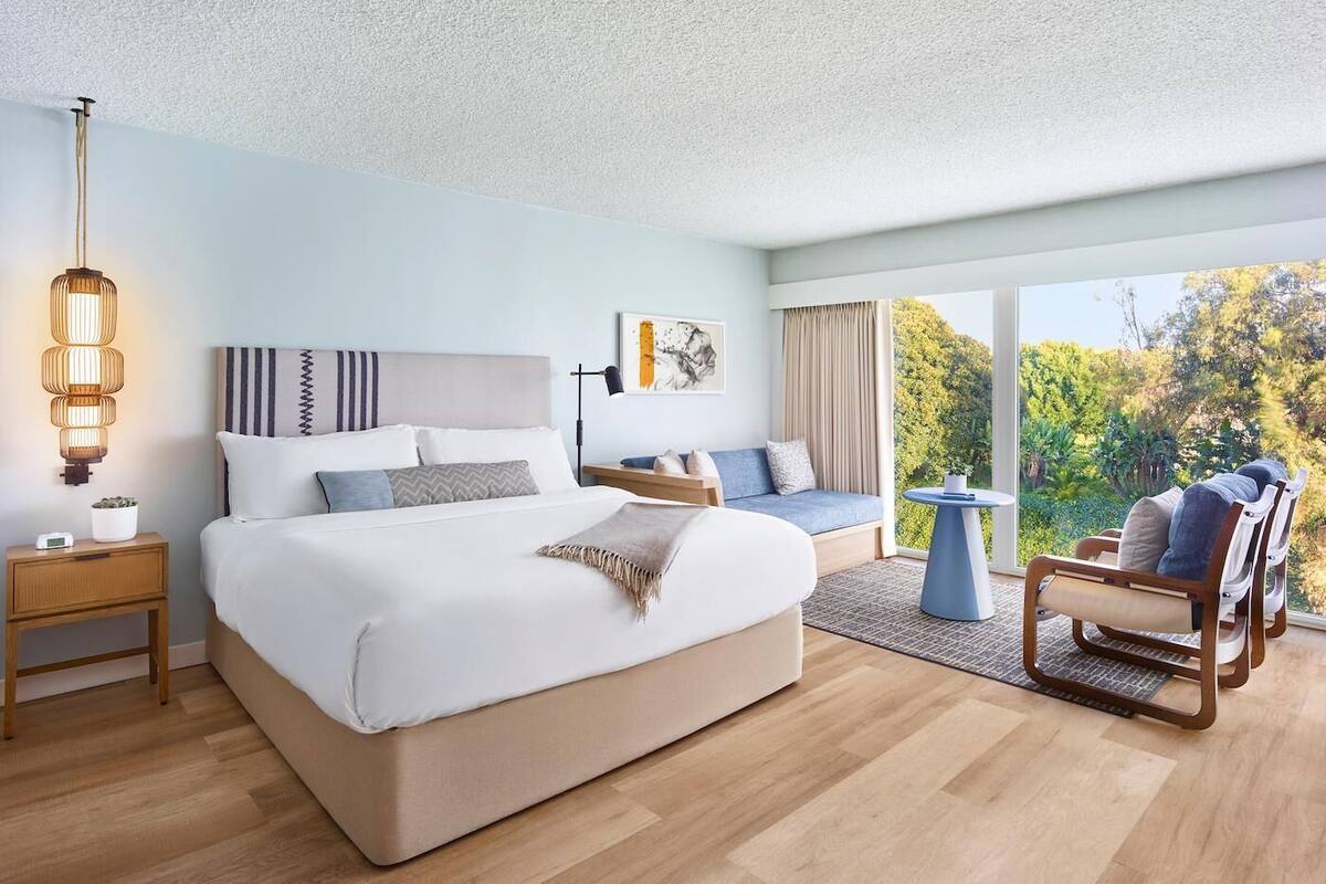 A modern bedroom with a large bed, bedside tables, a sofa, a chair, and a view of greenery.