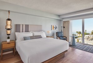 Habitación de hotel con una cama grande, mesitas de noche, una silla, una lámpara y vistas a la playa.