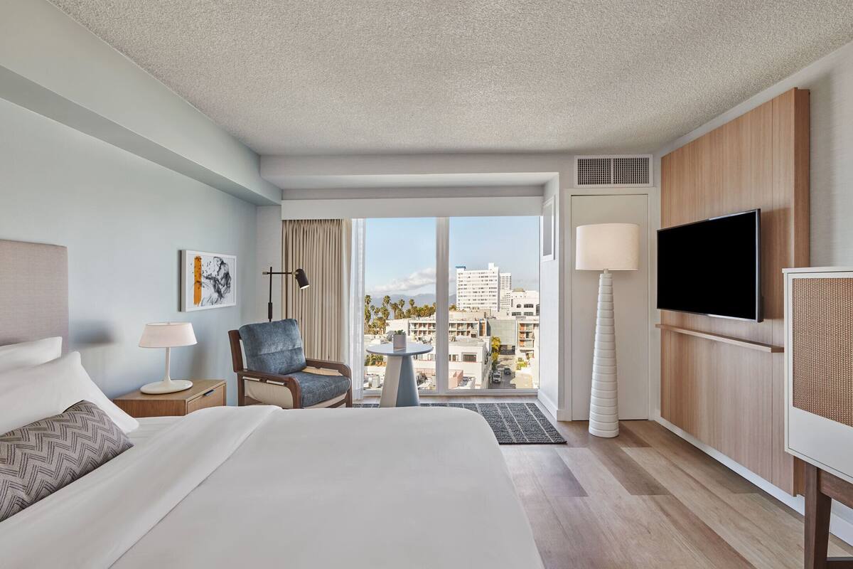 A hotel room with a bed, a chair, a TV, a lamp, a small table, and a view of the city.