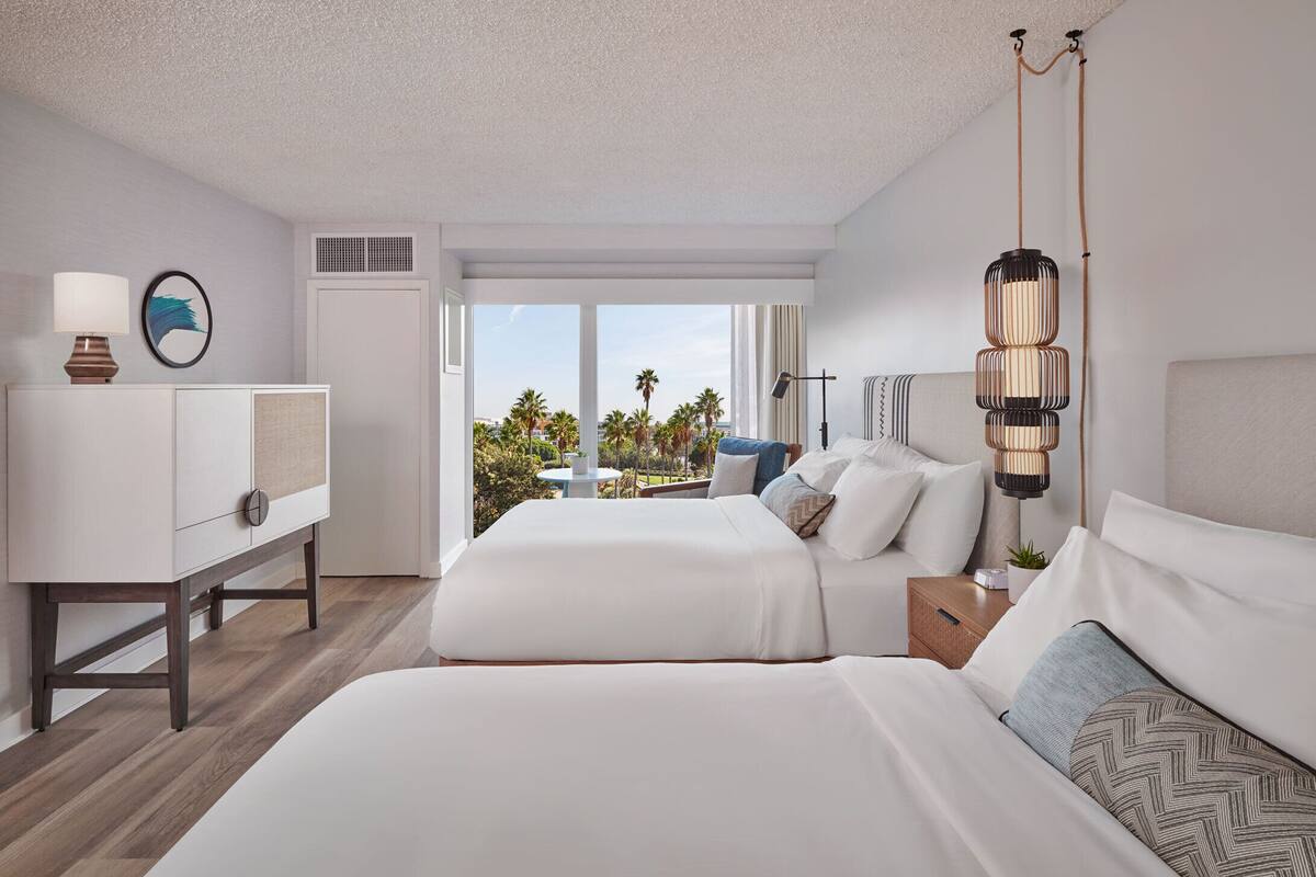 A hotel room with two beds, a desk, a lamp, and a view of palm trees.