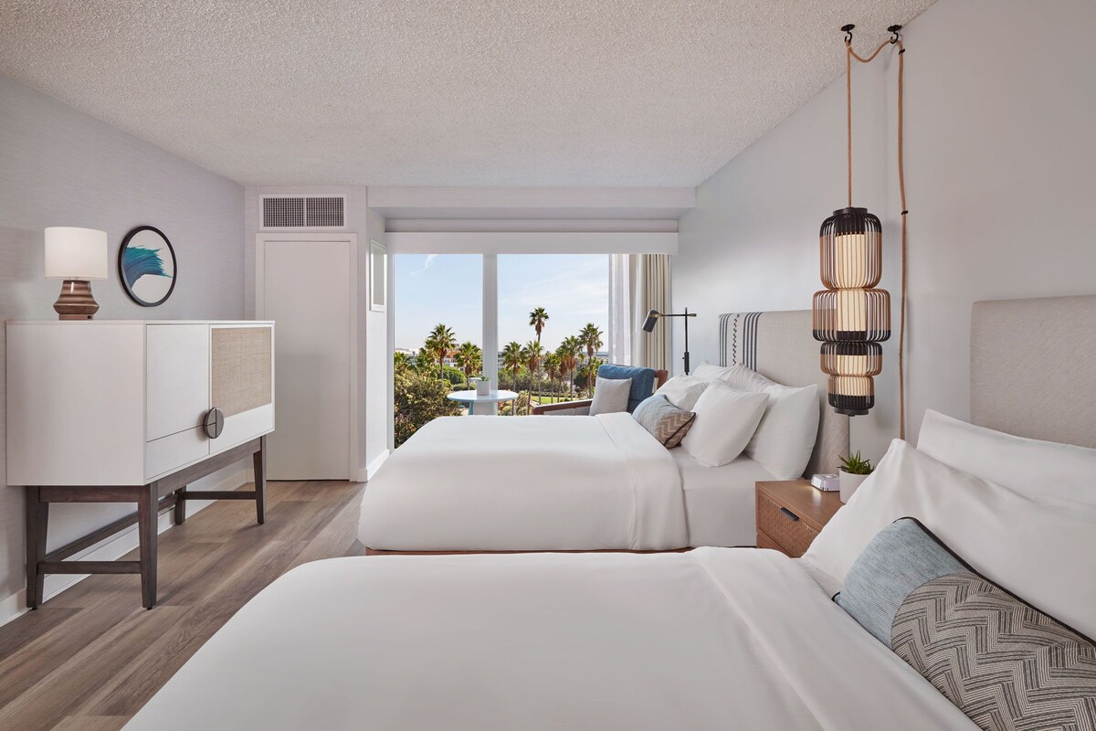 A hotel room with two beds, a desk, a lamp, and a view of palm trees.