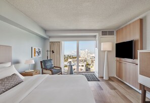 A hotel room with a bed, a chair, a TV, a lamp, a small table, and a view of the city.