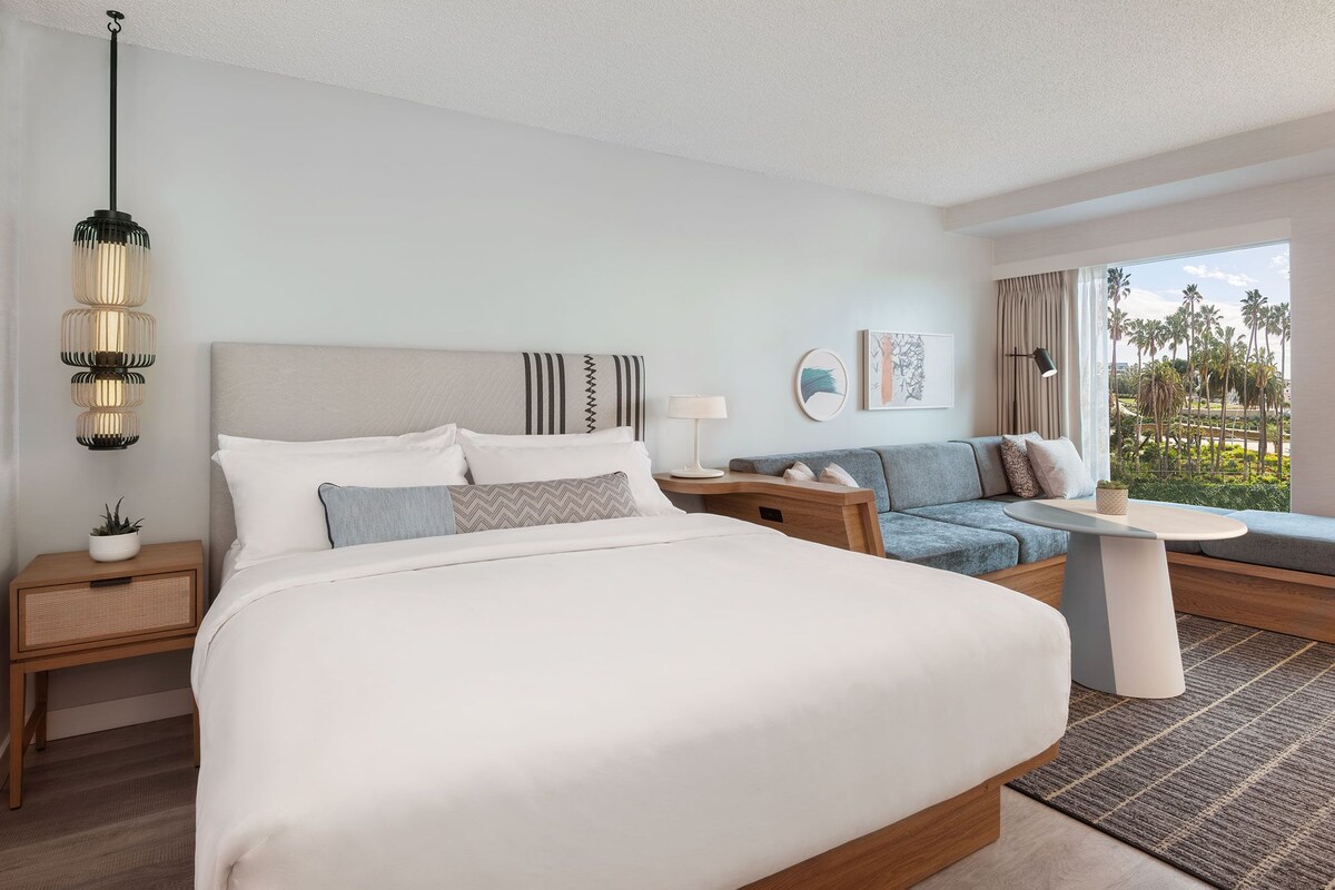 A modern hotel room with a large bed, a sofa, a small table, and a view of palm trees.