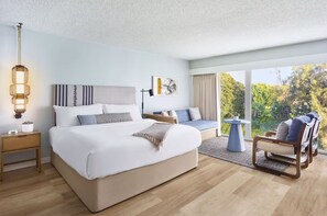 A modern bedroom with a large bed, bedside tables, a chair, and a view of greenery. - The Pierside (Santa Monica)