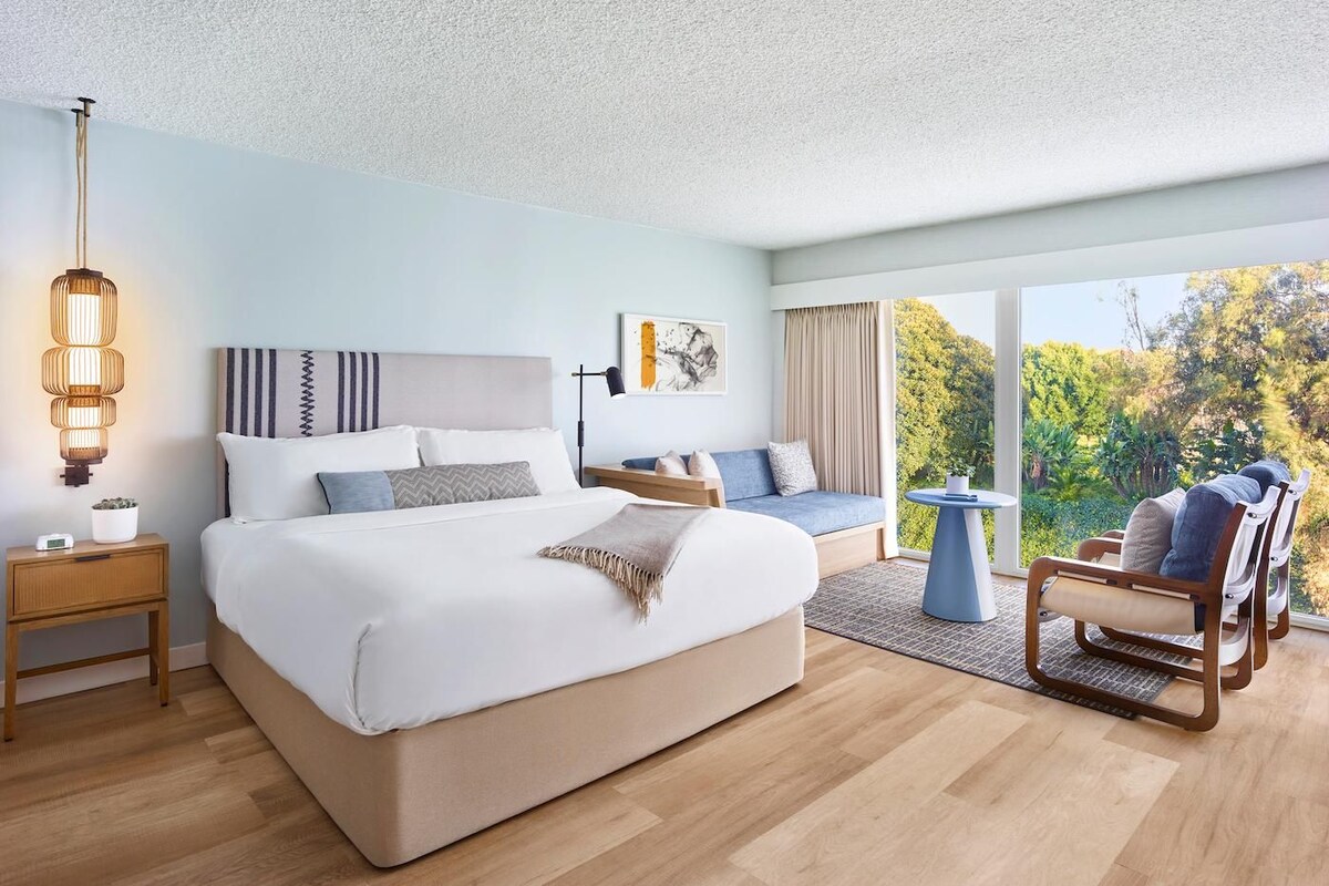A modern bedroom with a large bed, bedside tables, a sofa, a chair, and a view of greenery.