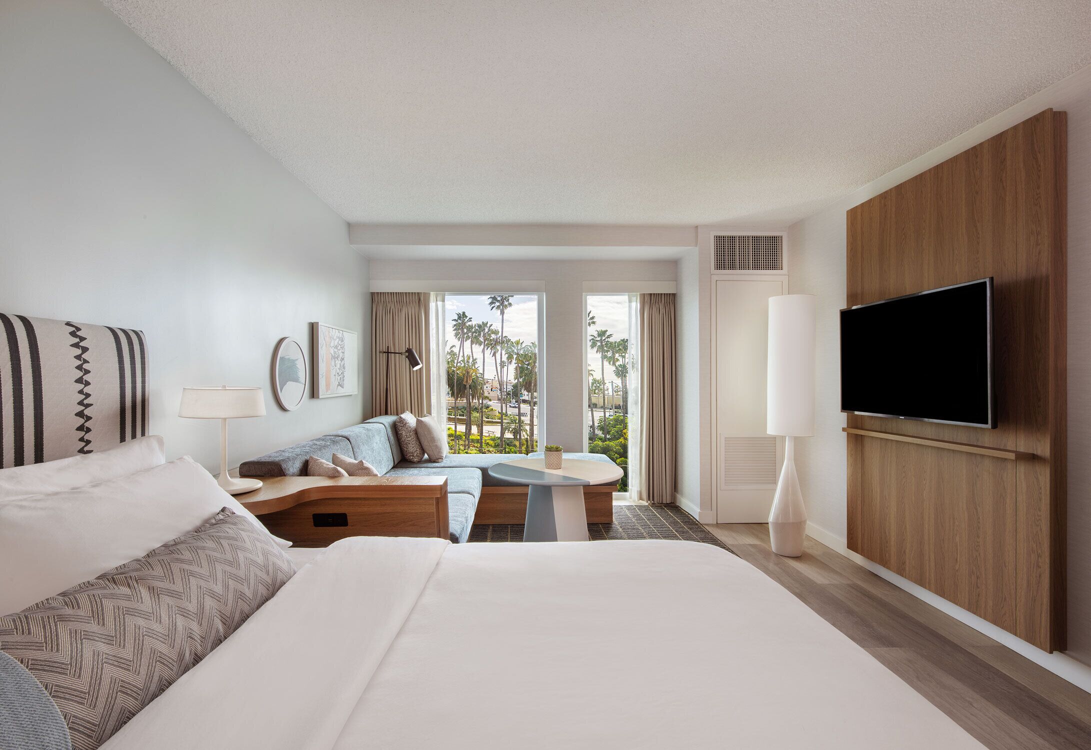 A hotel room with a bed, a sofa, a small table, a TV, and a large window with a view of palm trees.