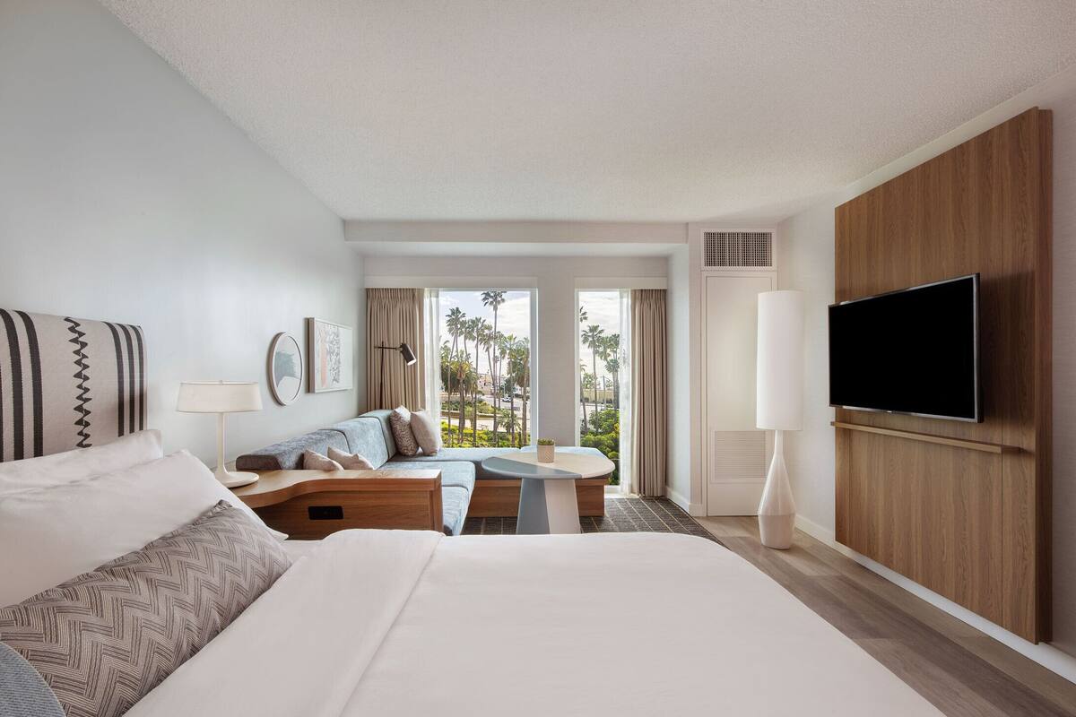 A hotel room with a bed, a sofa, a small table, a TV, and a large window with a view of palm trees.