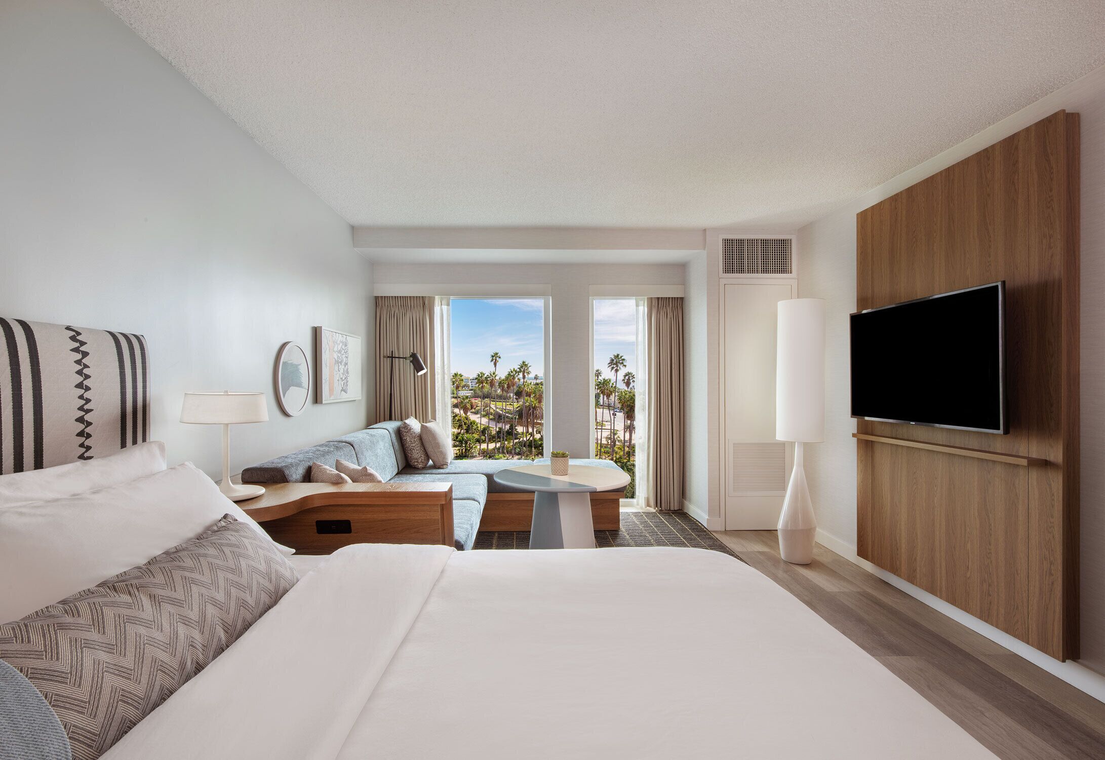 A hotel room with a large bed, a sofa, a small table, a TV, and a view of palm trees.