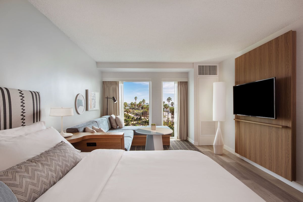 A hotel room with a large bed, a sofa, a small table, a TV, and a view of palm trees.