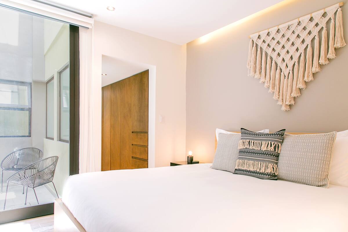 Ein modernes Schlafzimmer mit einem großen Bett, einer Wanddekoration aus Makramee und Blick auf eine Holztür.