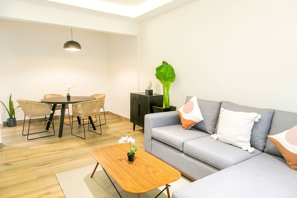 Ein modernes Wohnzimmer mit grauem Sofa, Couchtisch aus Holz, Korbstühlen, einem Esstisch und einem Blumentopf.