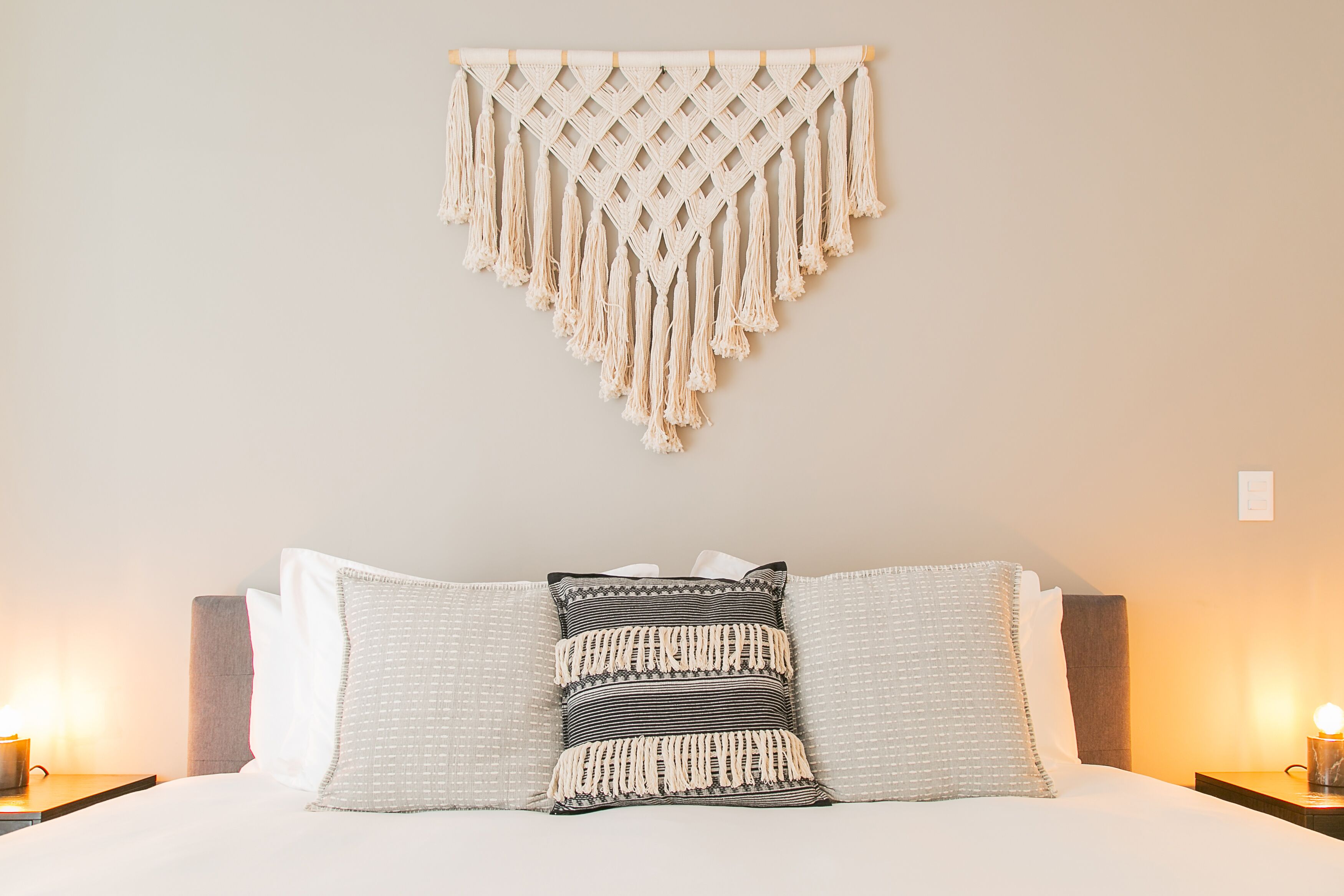 A neatly made bed with white and striped pillows, a macrame wall hanging, and bedside lamps.
