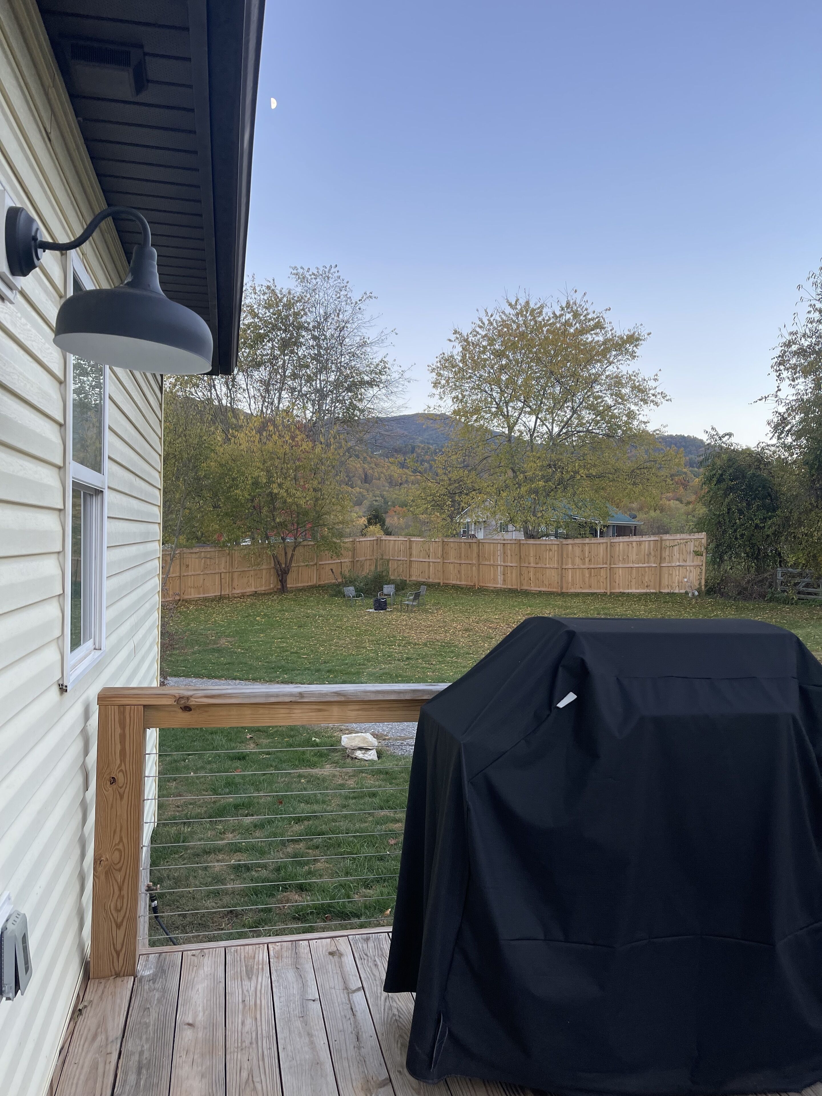 En terrasse med grill, et træhegn og udsigt over en baghave med træer og en fjerntliggende bakke.