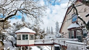 Een met sneeuw bedekte houten hut met een klein balkon en een terras dat naar een huis leidt.