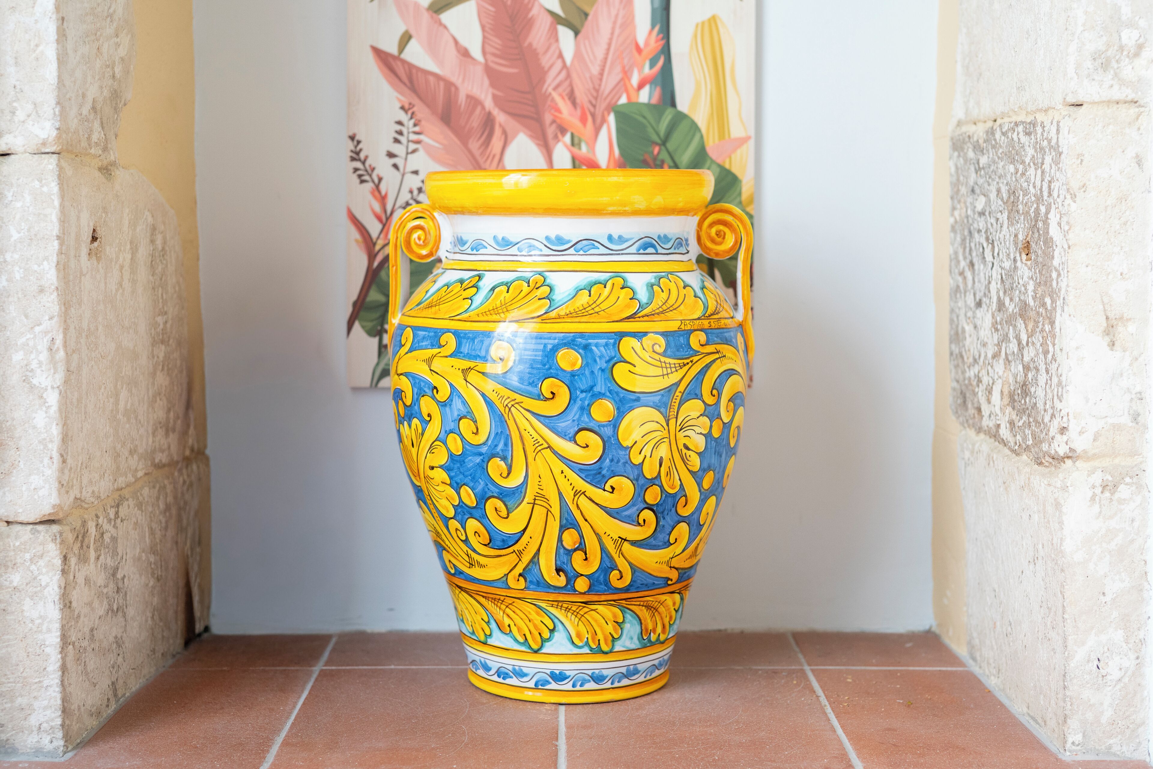 A large ceramic vase with yellow and blue patterns, placed on a tiled floor against a wall with a floral painting.