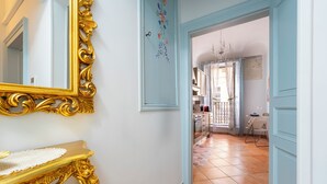 A hallway with a golden ornate mirror, a white console table, and a view into a kitchen with a tiled floor and a window with curtains.