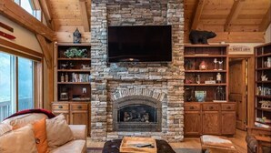 Ein gemütliches Wohnzimmer mit Kamin aus Stein, einem flachbildfernseher darüber und Holzböden mit verschiedenen Gegenständen.