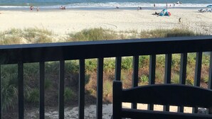 A beach view from a balcony with a chair, sandy beach, ocean, and a few people.