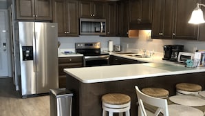 A modern kitchen with dark wooden cabinets, stainless steel appliances, a central island with stools, and a trash can.