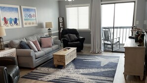 A spacious living room with a grey sofa, a black leather chair, a wooden coffee table, and a large window with blinds.