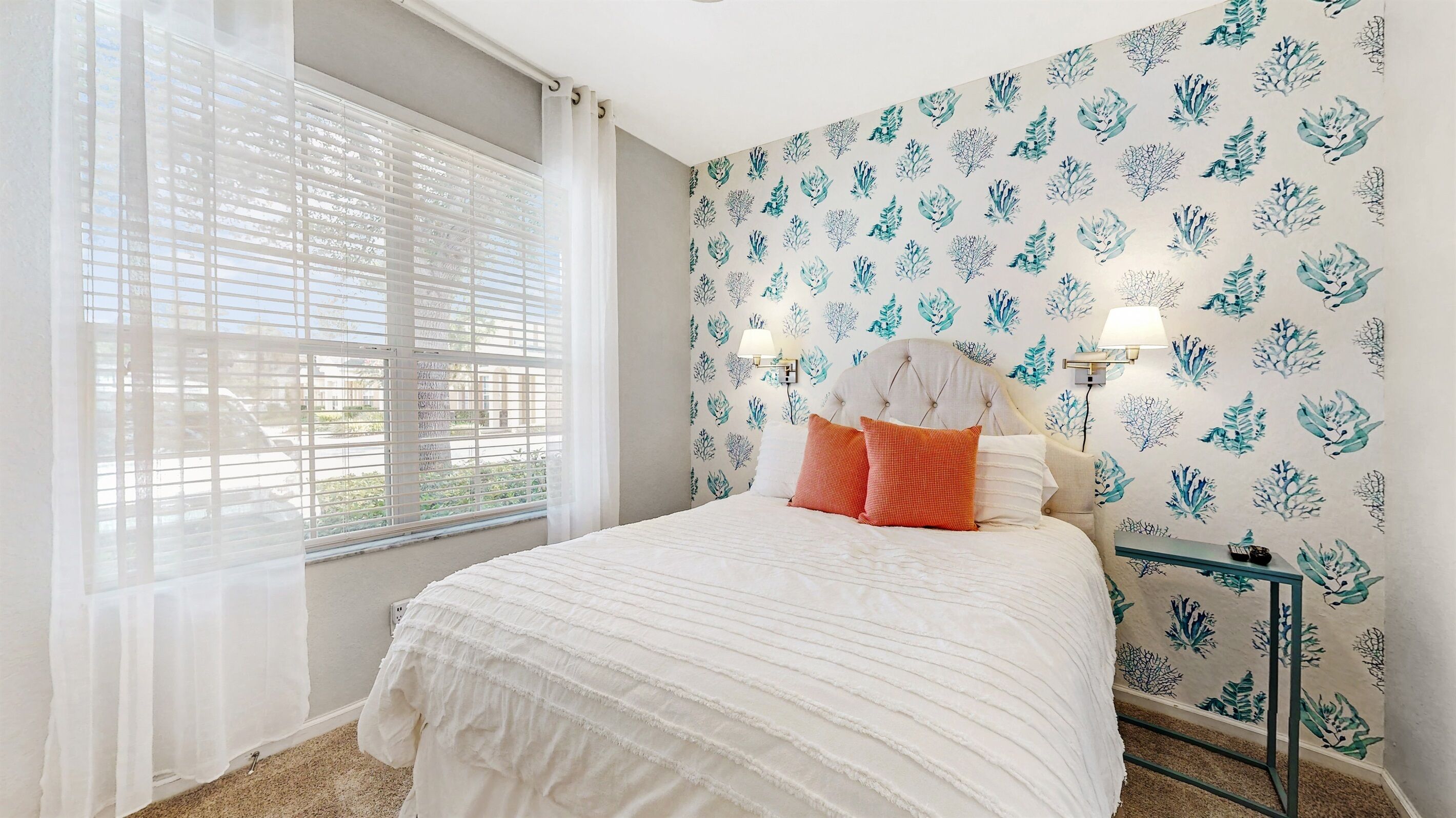 A bedroom with a bed, a window with blinds, a wall with a leaf pattern, and a bedside table with a lamp.