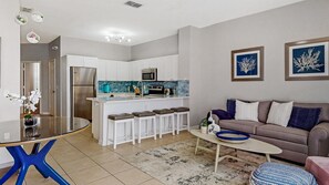 Una cocina moderna con armarios blancos, un refrigerador de acero inoxidable y un salpicadero de azulejos azules. Se observa un comedor con una mesa redonda y sillas azules. Se ve también una sala de estar con un sofá marrón y cojines estampados.