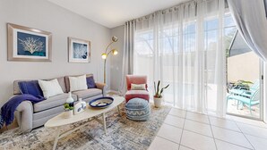 A modern living room with a grey sofa, a coffee table, and a pink armchair. There are two framed artworks on the wall, a floor lamp, and a view of the outside through large windows. - 3bd/3bth, Private Pool & View, 5mi to Disney Parks (Kissimmee)