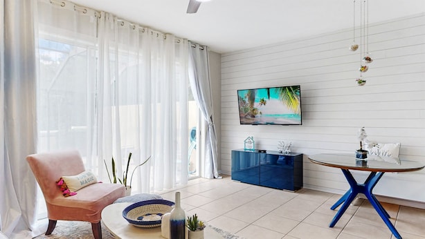 A modern living room with a pink armchair, a glass coffee table, a blue dining table, and a TV on the wall. - 3bd/3bth, Private Pool & View, 5mi to Disney Parks (Kissimmee)