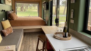 A cozy room with a bed, a small table with glasses and a bottle, a bench, and a view of a grassy area. - Marlo Hideaway (Marlo)