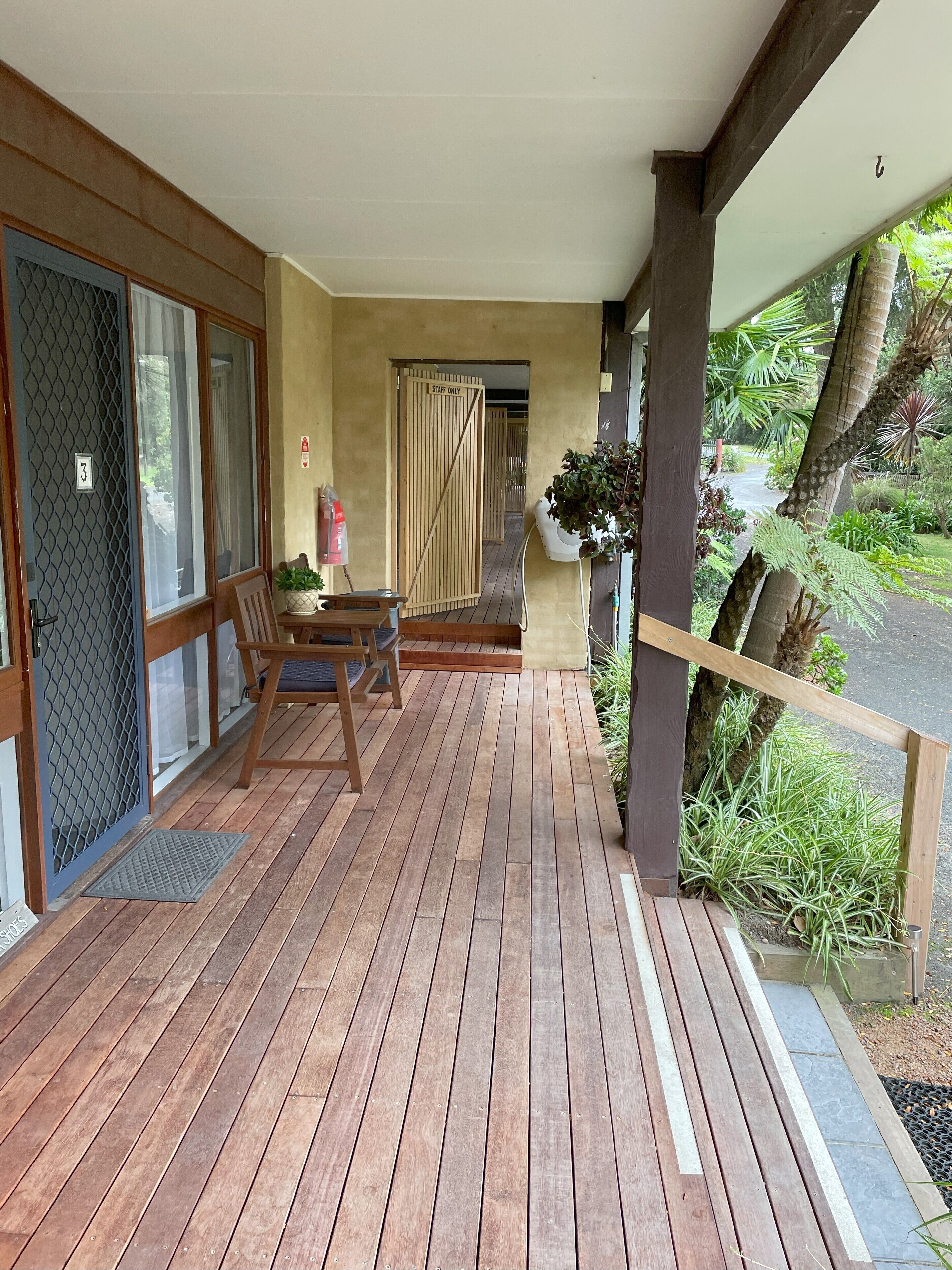 A wooden deck with a table and chairs, a door marked "Staff Only," and a view of a garden with plants and a palm tree.