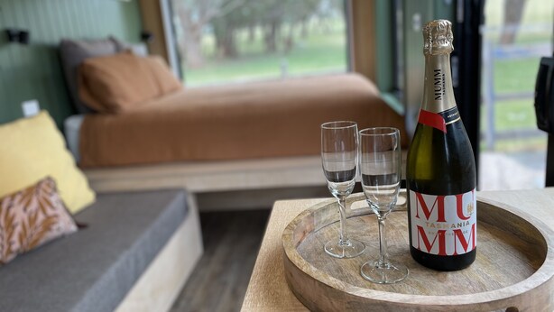 A bedroom with a bed, a wooden tray with a bottle of MUMM wine and two glasses, and a view of a green landscape through a window. - Marlo Hideaway (Marlo)