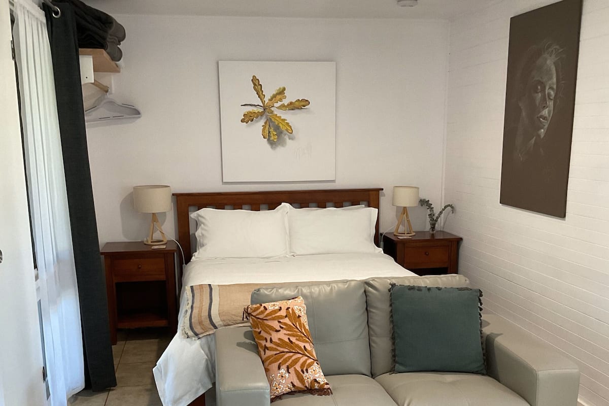 Une chambre à coucher avec un lit, un canapé gris, une table de chevet, une lampe fixée au mur et un ventilateur de plafond.