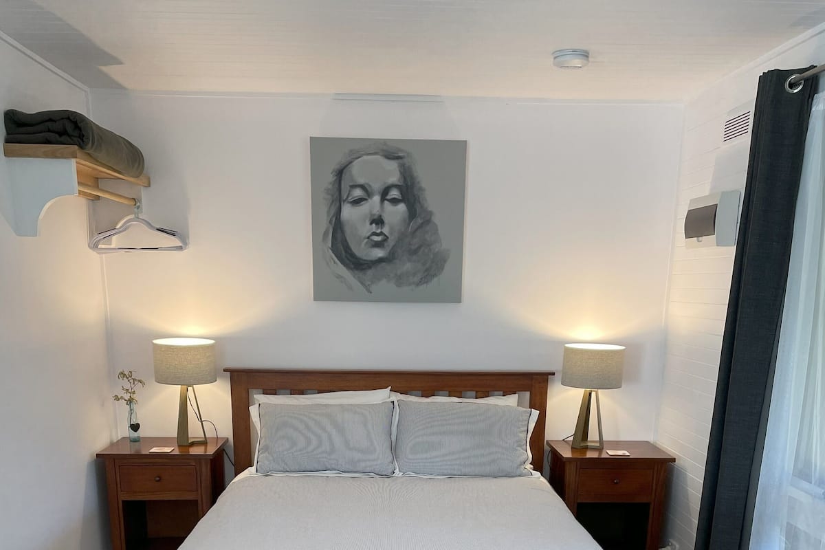 Une chambre à coucher avec un lit, deux tables de chevet avec des lampes, une tête de lit, une œuvre d’art accrochée au mur, une fenêtre avec des rideaux et un canapé de couleur claire avec un plaid.