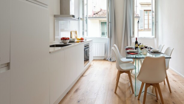 Uma cozinha moderna com armários brancos, uma área de jantar com mesa de vidro e vista para o exterior por grandes janelas.