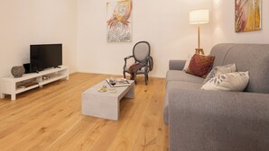 Uma sala de estar moderna com um sofá cinza, uma mesa de centro branca, uma TV sobre um suporte branco e um abajur.