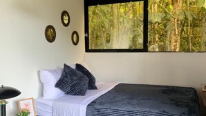 A bedroom with a bed, bedside table, lamp, and a window with a view of trees. - Hotel Selva Negra (Guatapé)