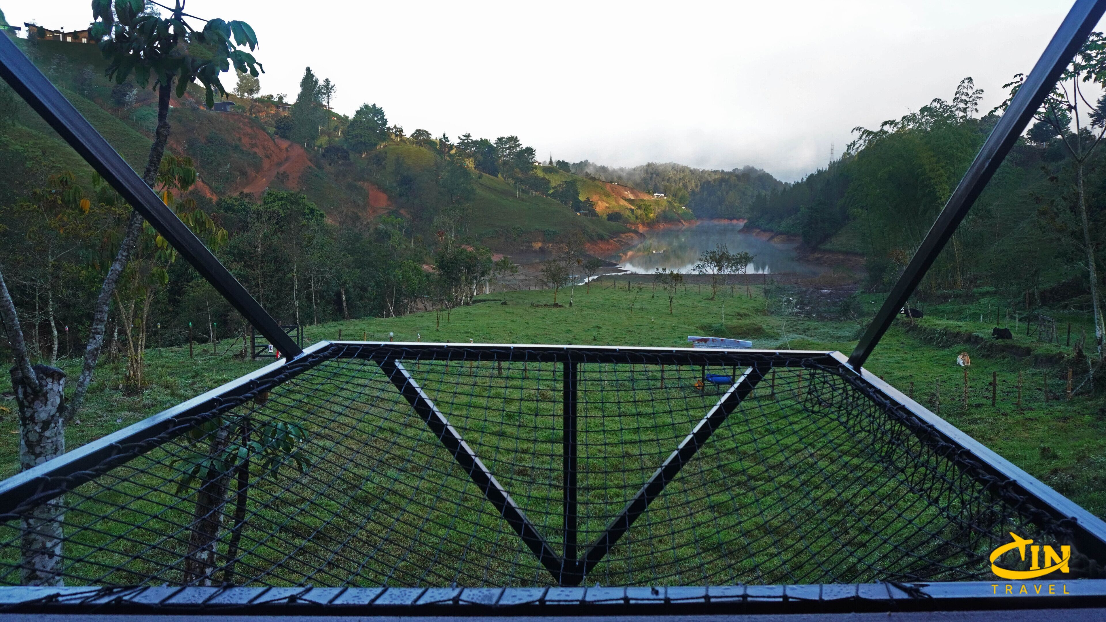 Vista desde un balcón con barandilla de metal, que da a un paisaje verde con una masa de agua, árboles y un edificio lejano.