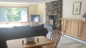 A living room with a stone fireplace, a television, a sofa, and a dining table with chairs. - Remodeled beautiful farmhouse with stunning lake view (Honeoye)