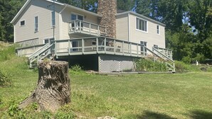 A two-story house with a stone chimney, a covered porch, and a large tree stump in the foreground. - Remodeled beautiful farmhouse with stunning lake view (Honeoye)