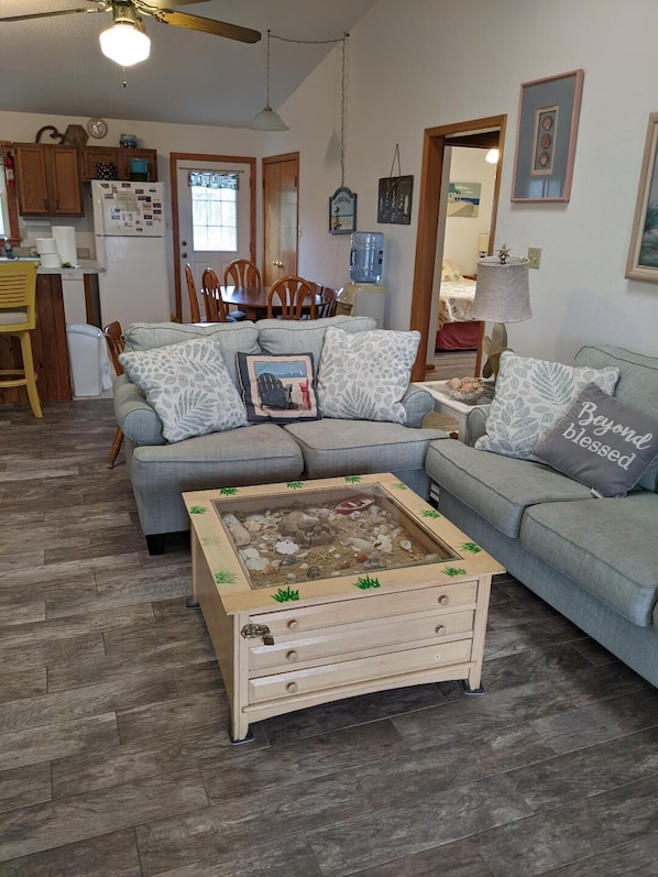 A cozy living room with a grey sofa, a wooden coffee table with a miniature beach scene, and a dining area in the background.