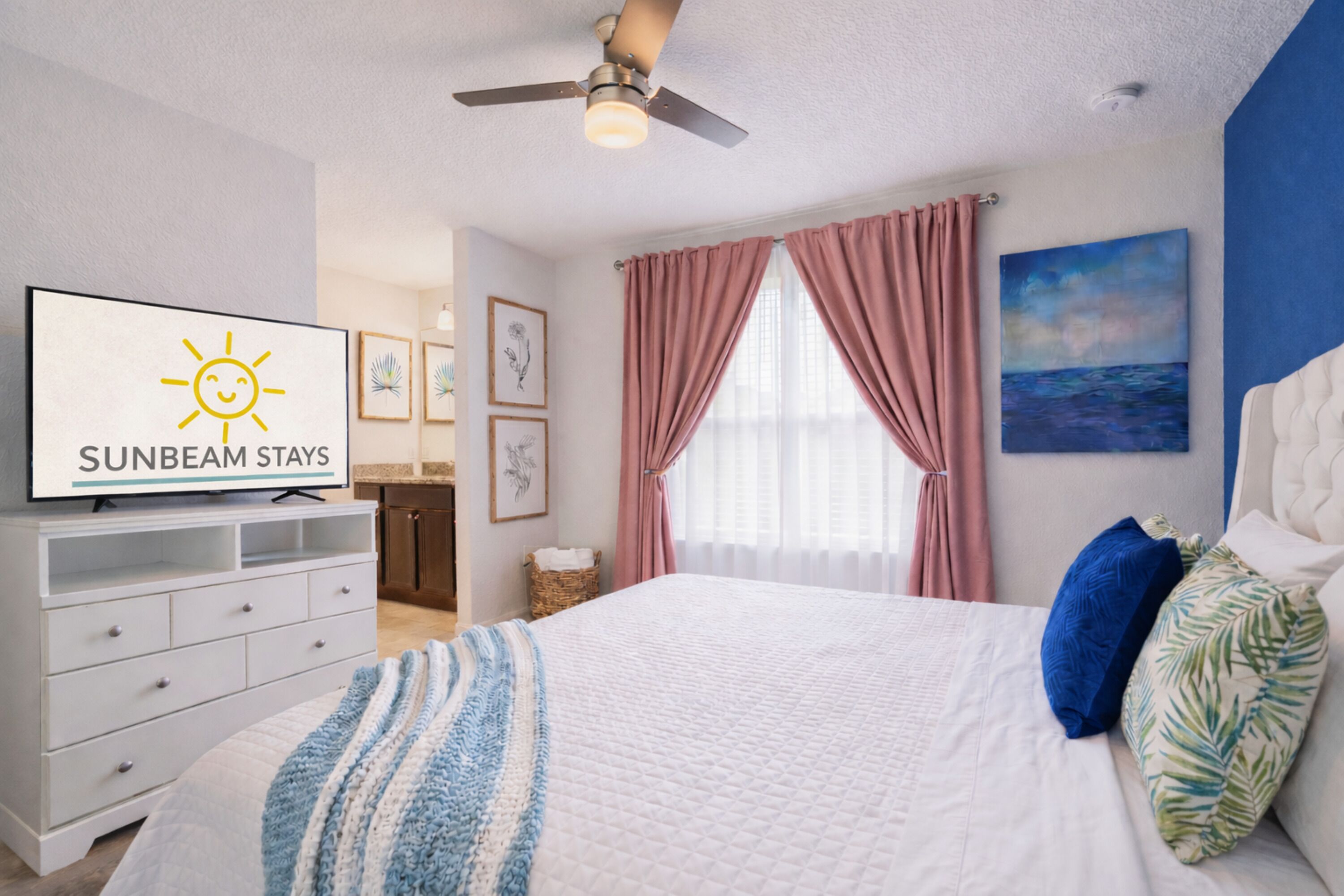 A bedroom with a bed, a television displaying 'SUNBEAM STAYS,' a ceiling fan, and a painting on the wall.