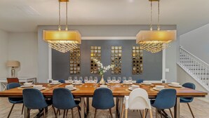 Uma sala de jantar com uma grande mesa de madeira, cadeiras azuis e duas lustres.