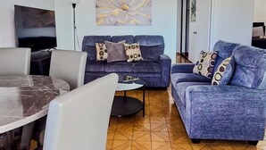 A living room with a blue sofa, a TV, a dining table, and a patterned floor. - Stunning 2 BR Private Apartment with Parking (Carolina)
