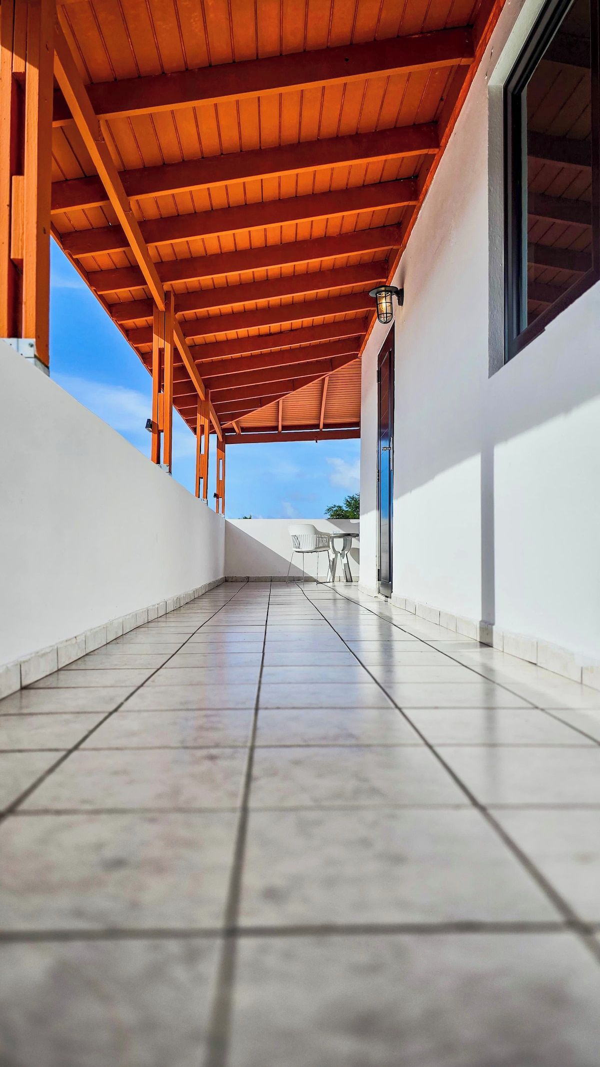 Un corredor techado al aire libre con suelo de baldosas, vigas de madera y vistas al cielo.