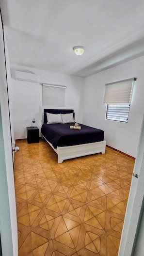 Un dormitorio con cama, mesita de noche, lámpara, ventana con persianas y luz de techo.