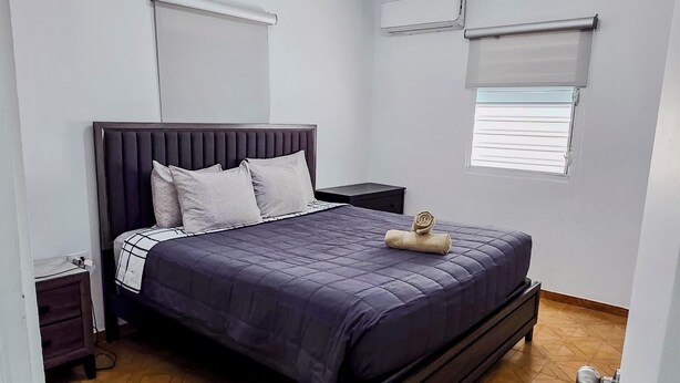 A bedroom with a bed, a nightstand, an air conditioning unit, and a window with blinds.
